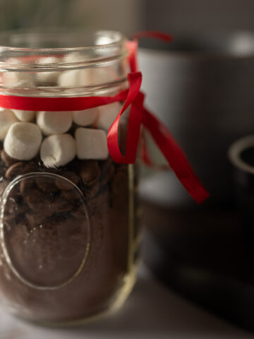 A jar of hot cocoa mix with two drinking mugs and marshmallows.