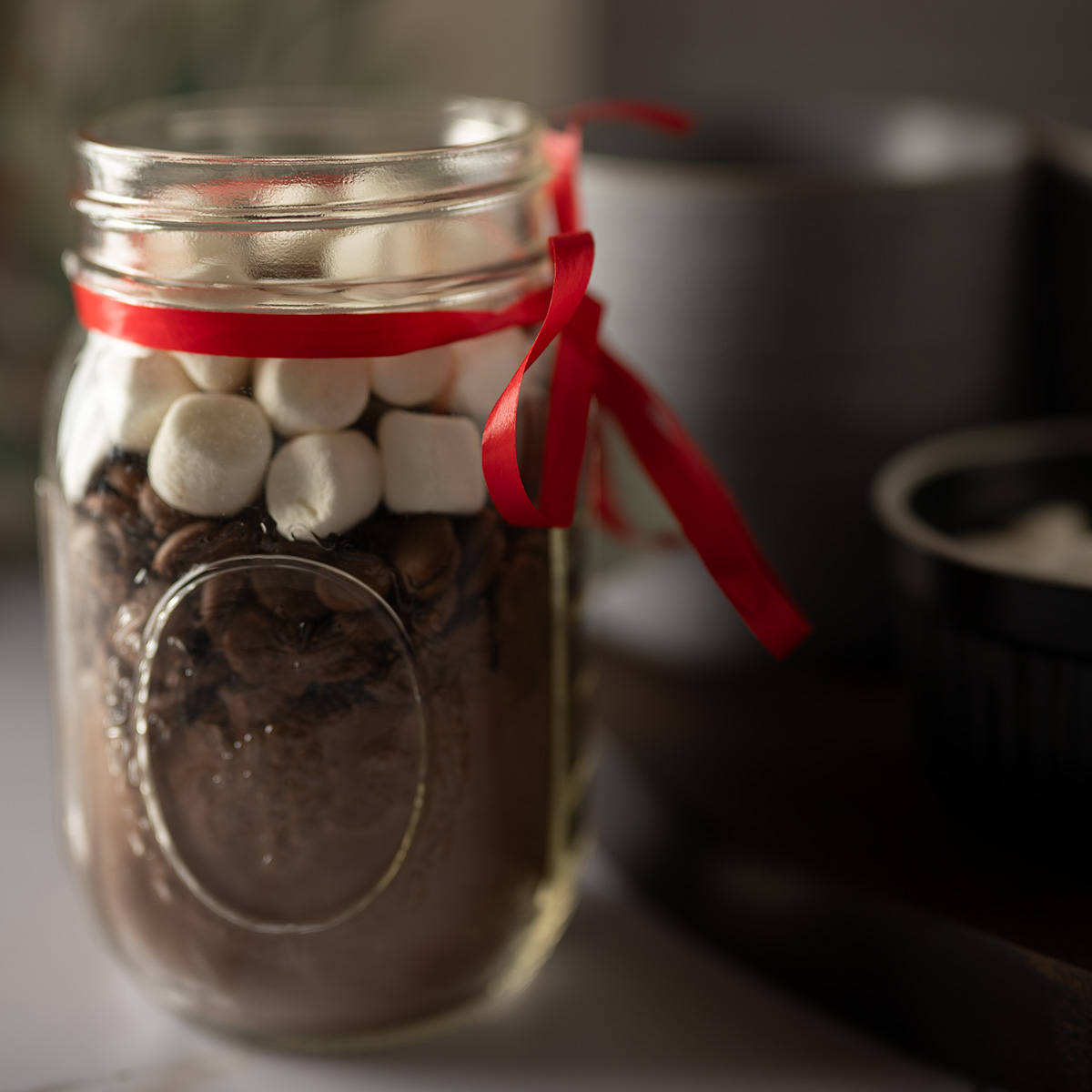 A jar of hot cocoa mix with two drinking mugs and marshmallows.